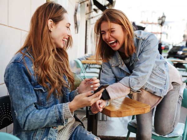 Two young female friends in casual stylish wear laughing while sitting at cafe in Madison, WI
